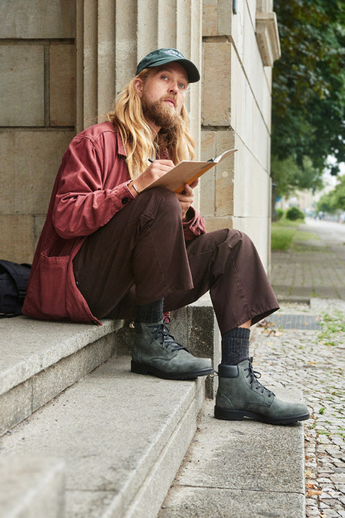 A person with long blond hair and a beard, wearing a green cap, red shirt, and brown pants, sits on stone steps outside, writing in a notebook and looking up thoughtfully. Trees and a sidewalk are visible in the background.
