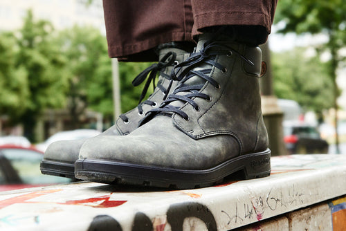 A close-up of a person wearing dark gray lace-up boots and brown pants, standing on a graffiti-covered surface outdoors, with blurred trees and cars in the background.