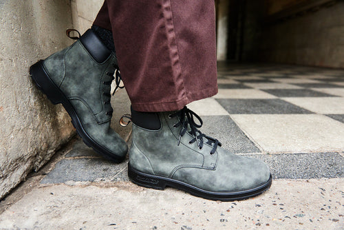 A person wearing green lace-up boots and brown pants stands on a black and white checkered floor next to a concrete wall.