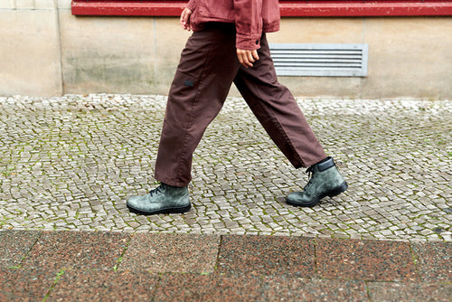 A person wearing brown pants, a reddish jacket, and gray boots walks on a cobblestone sidewalk past a stone wall with a red trim.