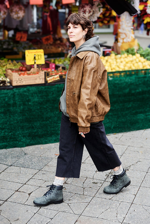 A person wearing a brown jacket, gray hoodie, wide black pants, and gray boots walks past a market stall with fruit and vegetables displayed in the background.
