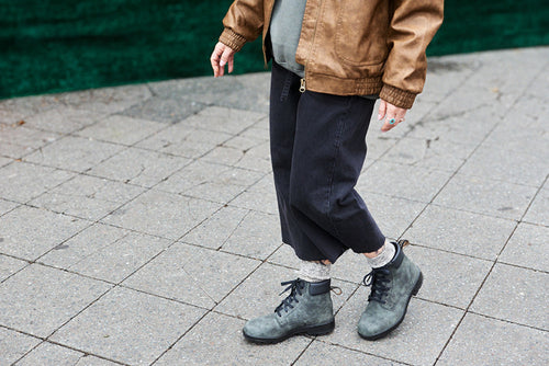 Person wearing a brown jacket, cropped black pants, grey socks, and grey lace-up boots walks on a stone-paved surface. Only the lower half of their body is visible. Green background in the upper part of the image.