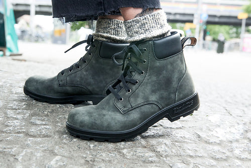 Close-up of a person wearing dark gray lace-up boots and light gray wool socks, standing on a textured stone surface outdoors. Only the lower legs and feet are visible.