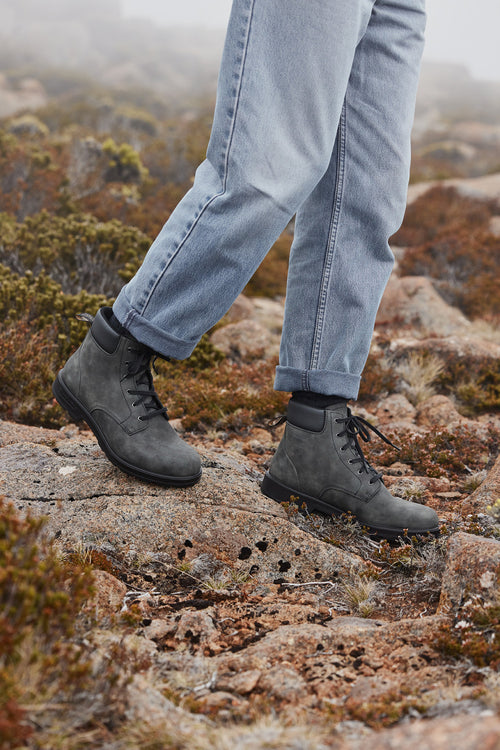 A person wearing cuffed light blue jeans and dark gray hiking boots walks on rocky, grassy terrain outdoors.