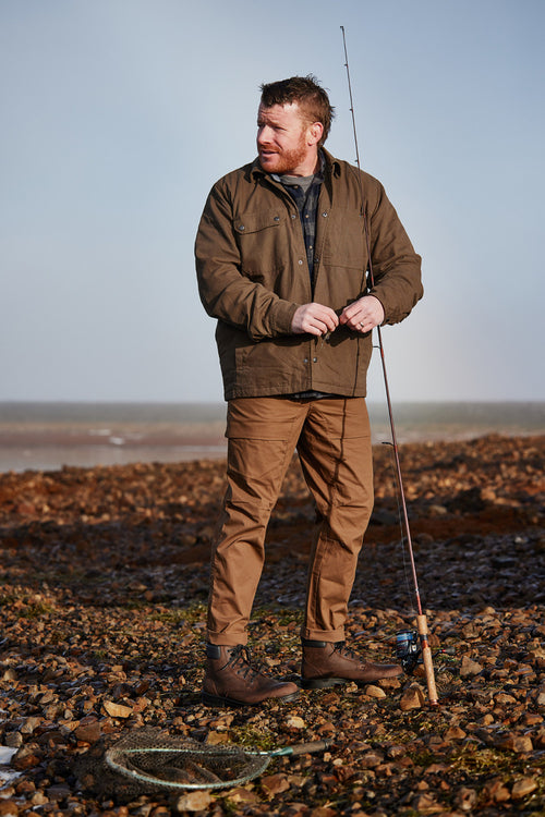 A man stands on a rocky shoreline holding a fishing rod, dressed in a brown jacket, tan pants, and boots, with a fishing net on the ground nearby under a cloudy sky.
