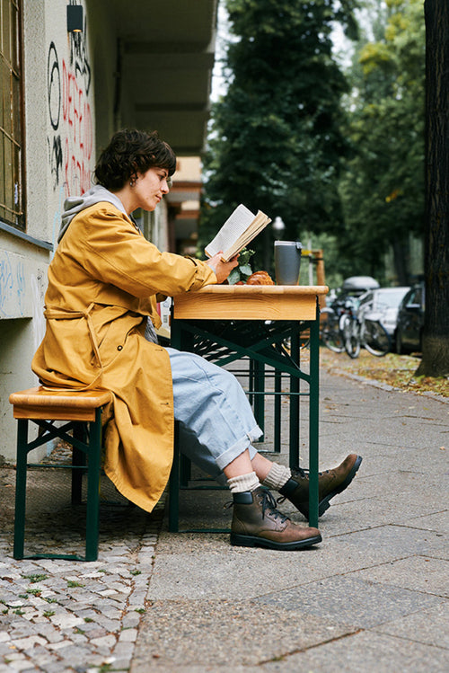 A person in a yellow coat sits at an outdoor table on a city sidewalk, reading a book. There’s a drink and pastry on the table, and bicycles are parked in the background among trees and buildings.