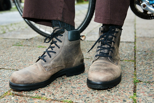 A person wearing dark brown pants and gray lace-up boots stands on a paved sidewalk next to a bicycle wheel.
