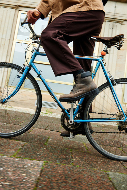 A person wearing brown pants and boots is riding a blue bicycle on a city sidewalk near a building with large windows. The photo is taken from a low angle, focusing on the bike and the rider’s legs.