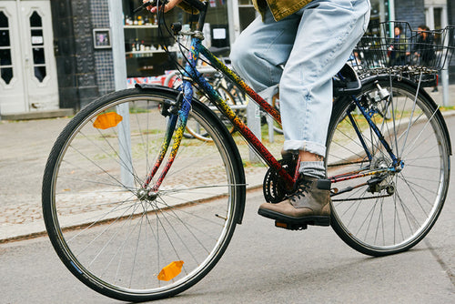 A person wearing rolled-up jeans and brown boots rides a colorful bicycle with a basket on the back along a city street.