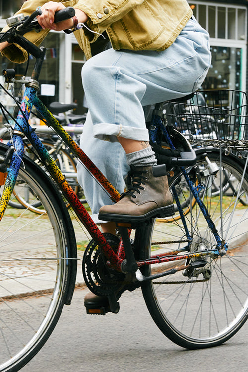A person wearing rolled-up jeans, gray socks, and brown boots is riding a colorful bicycle on a city street. Only their lower body and the side of the bike are visible.