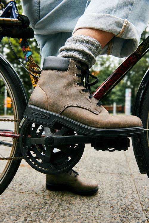 A person wearing rolled-up jeans, gray socks, and brown boots rests their foot on a bicycle pedal, with the bike frame and chain visible. The background is outdoors with blurred greenery.