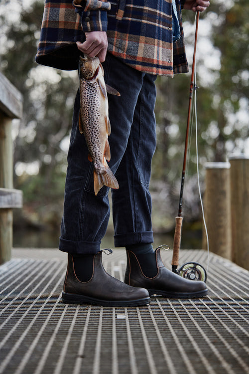 A person in dark pants, boots, and a plaid shirt stands on a dock, holding a fishing rod in one hand and a freshly caught trout in the other. The background shows blurred trees and water.