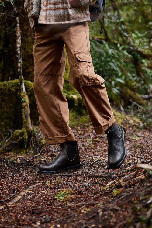 Person wearing brown cargo pants and black boots walks on a leaf-covered forest trail, surrounded by green moss and foliage. Only the lower half of the body is visible.