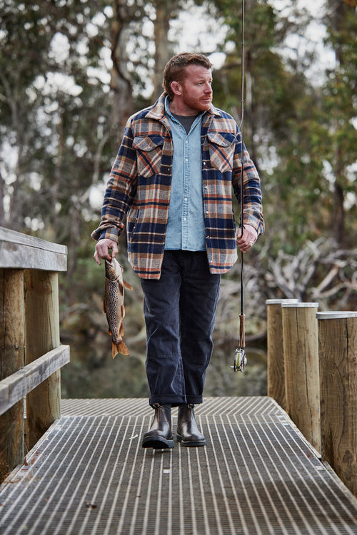 A man in a plaid jacket and boots stands on a dock holding a fishing rod in one hand and a fish in the other, with trees and water in the background.