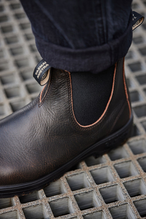 A close-up of a persons foot wearing a brown leather Chelsea boot with black elastic side panels and dark jeans, standing on a metal grid surface.