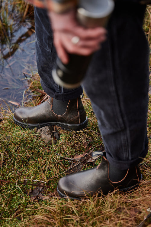 Person wearing brown leather boots stands on grassy ground near water, holding a metallic thermos. Only the lower half of the person is visible.