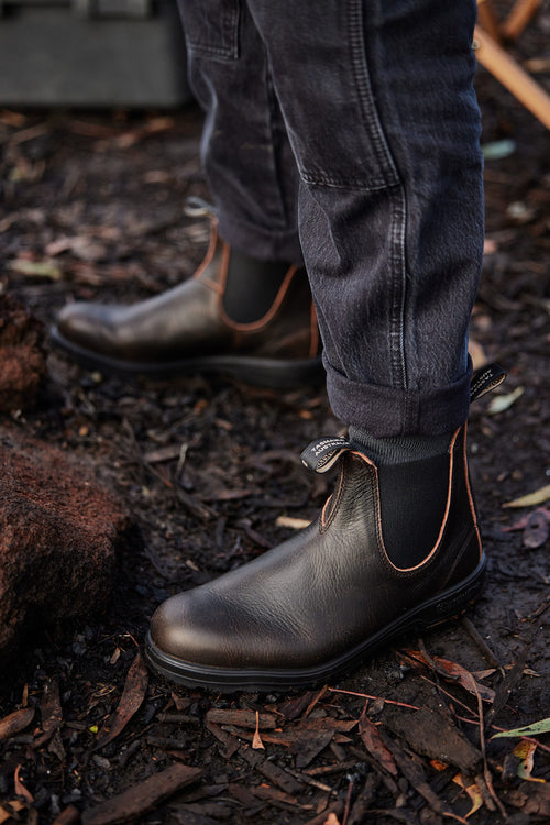 Person wearing dark leather boots and rolled-up black pants standing on soil covered with dried leaves and twigs.