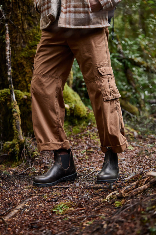 A person stands in a forest wearing brown cargo pants, a plaid jacket, and dark brown Chelsea boots. The ground is covered with damp leaves and moss, and green foliage surrounds the scene.
