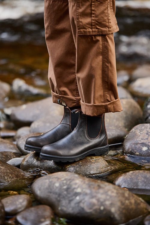 A person wearing brown cargo pants and black leather boots stands on wet rocks in a shallow stream.