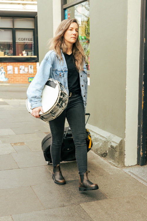 A woman with long hair, wearing a denim jacket and black boots, walks on a city sidewalk carrying a snare drum under her arm, with a black bag on the ground behind her.