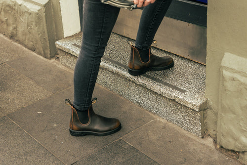 A person wearing black skinny jeans and dark brown ankle boots steps up onto a granite doorstep, holding a small bag. The scene is on a paved sidewalk near a building entrance.