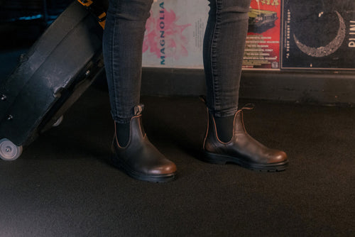Person wearing dark brown Chelsea boots and skinny jeans stands indoors on a dark carpeted floor, next to a wheeled suitcase. Colorful posters are visible on the wall in the background.