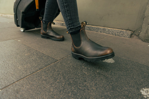 A person wearing dark brown Chelsea boots and black jeans walks on a sidewalk, pulling a black wheeled suitcase behind them.