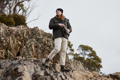 A person with long hair, wearing a dark jacket, beige pants, boots, and a black beanie, stands on rocky terrain holding binoculars. Trees and cloudy sky are visible in the background.