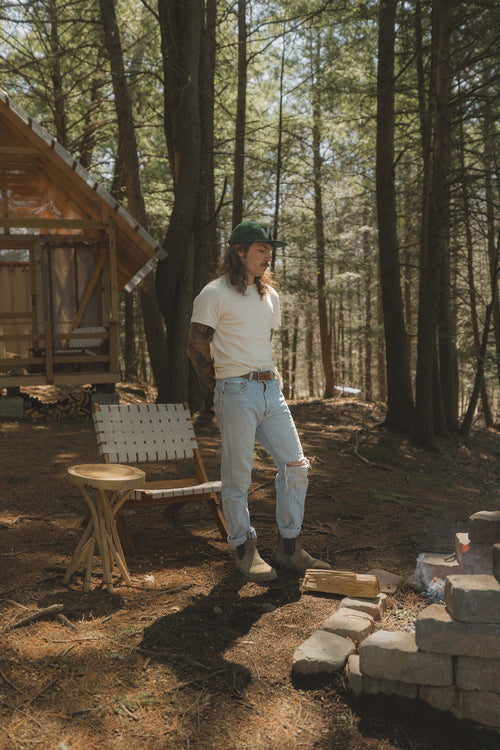 A person with long hair and tattoos, wearing a green hat, white t-shirt, and ripped jeans, stands near a fire pit in a wooded area beside a wooden cabin and a folding chair. Sunlight filters through the trees.