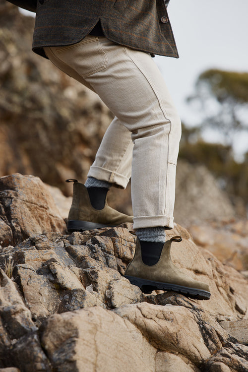 A person wearing light-colored pants and olive green Chelsea boots is climbing over rocky terrain outdoors. Only their lower body and legs are visible.