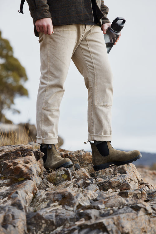 A person wearing beige pants and green boots stands on rocky ground outdoors, holding a water bottle. The persons head is not visible, and there are trees and a cloudy sky in the background.