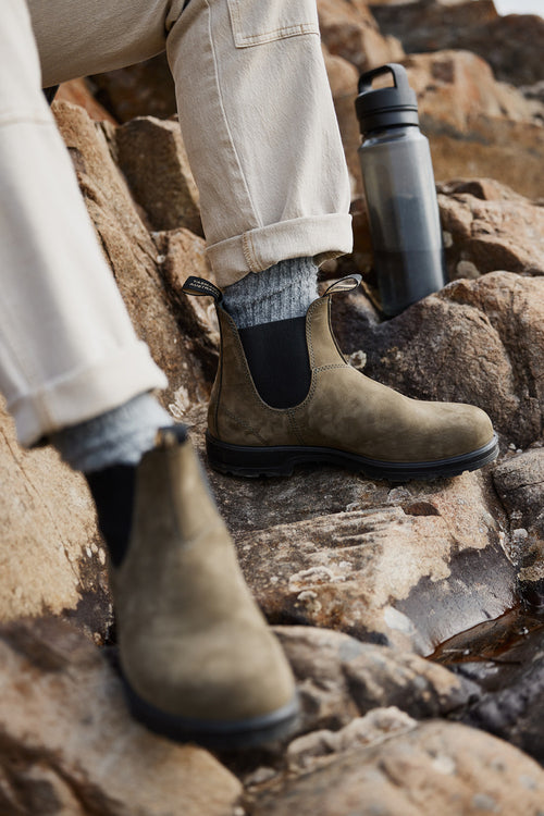 A person wearing light-colored pants, gray socks, and brown Chelsea boots sits on rocky terrain next to a black water bottle.