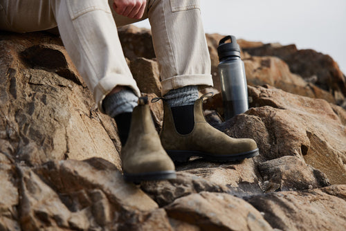 A person wearing tan boots, gray socks, and light pants sits on rocky terrain next to a reusable water bottle. Only their lower legs and feet are visible.