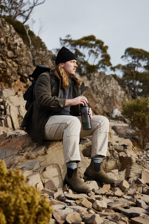A man with long hair and a beard, wearing a beanie and outdoor clothing, sits on a rock in a rocky, mountainous area, holding a thermos and looking into the distance. Bushes and trees are in the background.