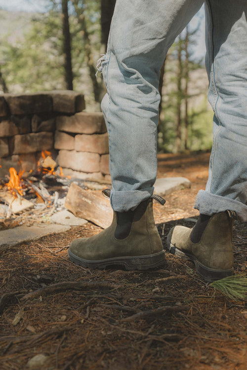 A person wearing light blue jeans and brown boots stands near a campfire in a forested area, with stacked bricks and trees visible in the background.