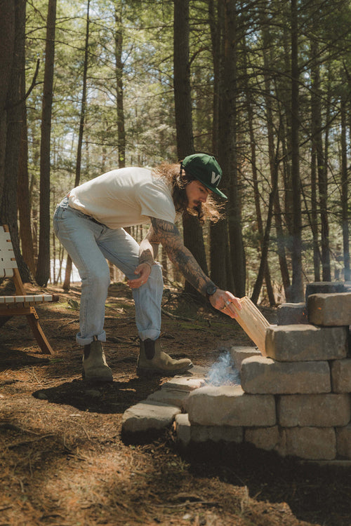 A person with long hair, tattoos, and a green cap is placing a piece of wood on a campfire built with stacked stones in a forested area. Sunlight filters through tall trees, and there is a wooden chair nearby.