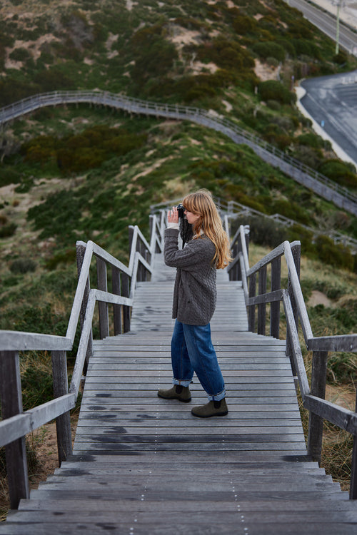 A person with long hair, wearing a grey sweater and blue jeans, stands on a wooden stairway outdoors, looking through binoculars at a grassy landscape with winding paths.