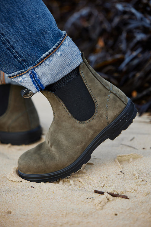 A person wearing cuffed blue jeans and dark socks steps on sandy ground, showcasing an olive green suede Chelsea boot with black elastic side panels.