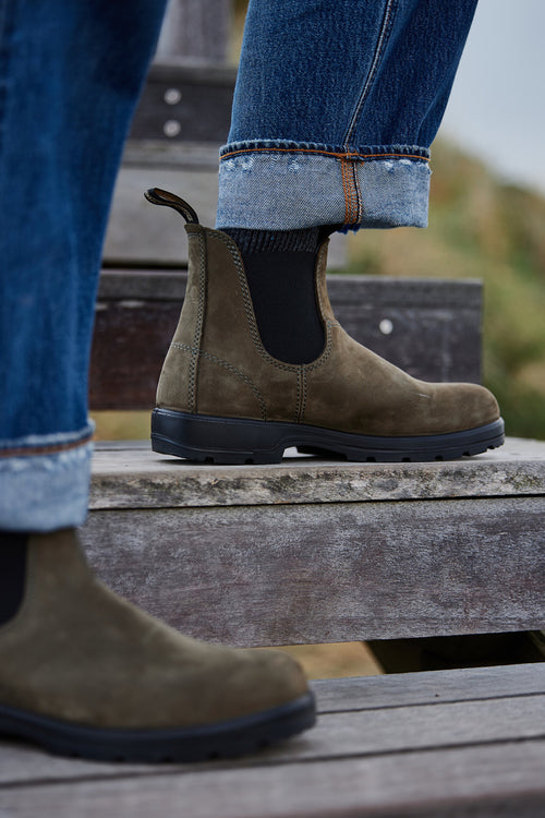 A person wearing dark brown Chelsea boots and cuffed blue jeans stands on wooden steps outdoors. The focus is on the boots and lower legs.