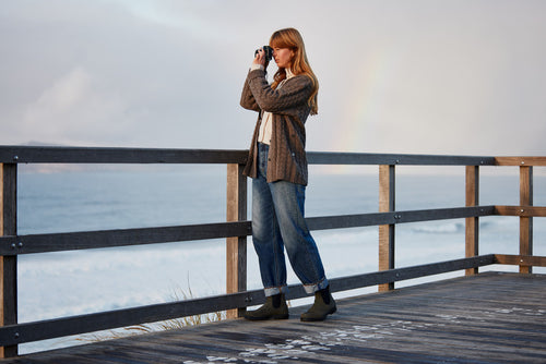 A woman with long hair stands on a wooden deck overlooking the ocean, using binoculars. She is wearing a brown jacket, jeans, and boots. The sky is cloudy with a faint rainbow visible in the background.