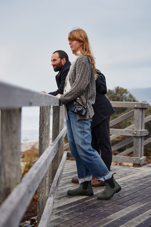 A woman and a man stand on a wooden boardwalk overlooking the water. The woman has a camera around her neck and is wearing jeans, boots, and a knit sweater. Both appear relaxed and are gazing into the distance.