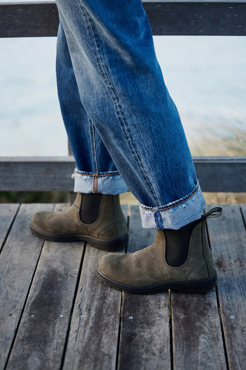 Person standing on a wooden deck wearing cuffed blue jeans and brown Chelsea boots, with a railing and blurred outdoor background visible.