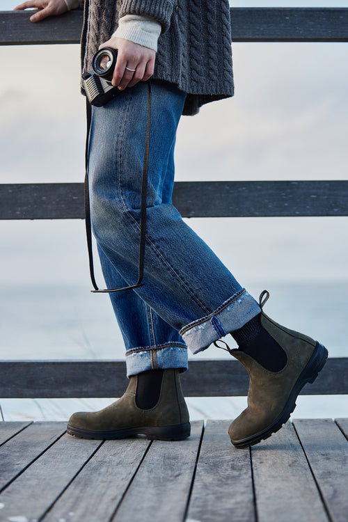 A person in rolled-up jeans and brown boots stands on a wooden deck by the water, holding a camera. The upper body is out of frame, and the background is soft and cloudy.
