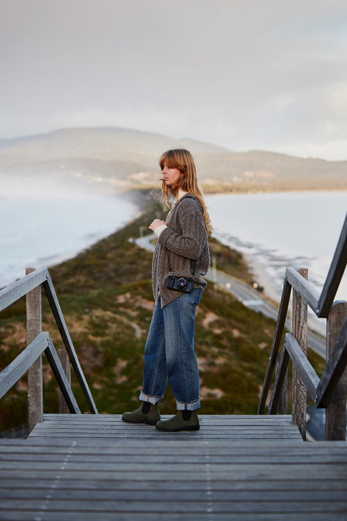 A woman stands on wooden stairs overlooking a scenic coastal landscape, with ocean on both sides, mountains in the background, and a camera hanging from her neck.