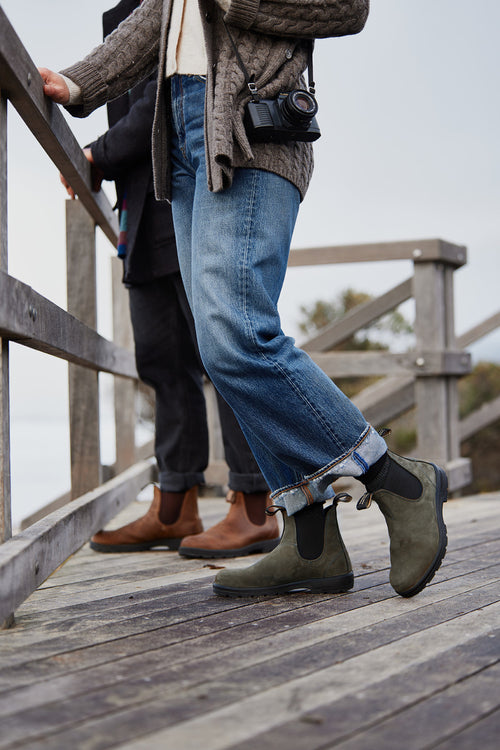 Two people stand on a wooden deck outdoors, one wearing green Chelsea boots, blue jeans, a cardigan, and a camera around their neck; the other wears brown boots and dark pants.