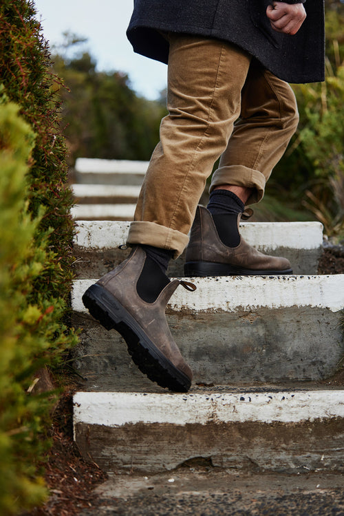 A person wearing brown pants and dark brown boots walks up concrete steps outdoors, surrounded by green bushes. Only their legs and part of a dark coat are visible.