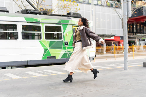A woman wearing a brown jacket and cream skirt walks confidently across a city street in front of a green and white tram.