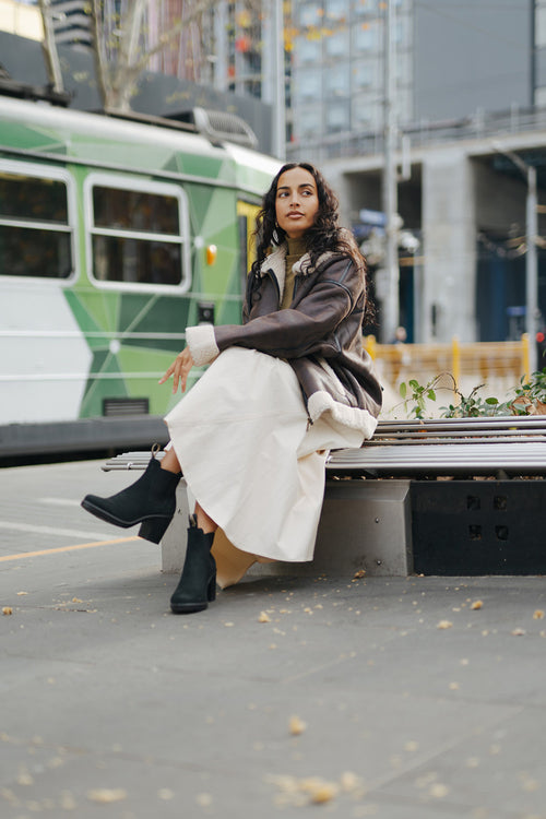 A woman with long curly hair sits on a bench outdoors, wearing a brown jacket, beige skirt, and black boots. A green and white tram passes by in the background on a city street.