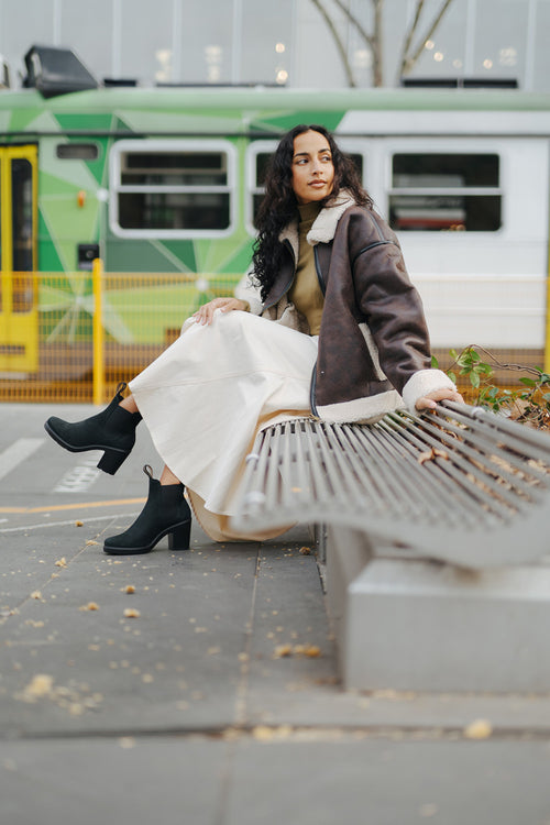 A woman wearing a shearling jacket, long cream skirt, and black heeled boots sits on a modern bench outdoors, with a green tram passing by in the background.