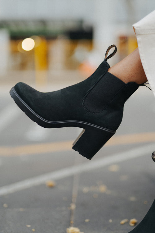A close-up of a person’s foot wearing a black suede heeled ankle boot with a loop pull tab, paired with cream-colored pants, photographed outdoors on a city street.